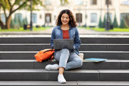 Photo for Joyful young indian woman student studying online, using laptop computer outdoors, sitting on stairs at college campus, doing homework, copy space. E-learning concept - Royalty Free Image