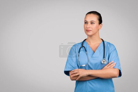 Photo for Confident young female nurse in teal medical scrubs with stethoscope around the neck, arms folded, standing against grey studio background, free space - Royalty Free Image