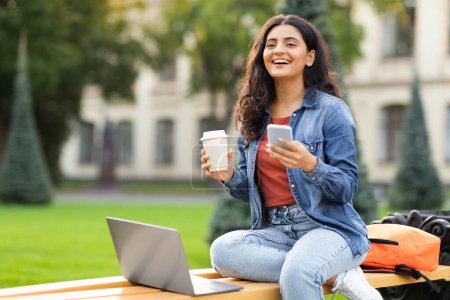 Téléchargez les photos : Joyeux beau jeune étudiante indienne travaillant sur le projet, assis sur un banc dans une zone urbaine de la ville, utilisant un ordinateur portable à l'extérieur, vérifiant smartphone, buvant du café, espace libre - en image libre de droit