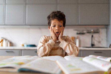 Foto de Chico negro asombrado que parece sorprendido mientras estudia con libros de texto en casa, estresado niño afroamericano masculino sentirse abrumado con la tarea o proyectos escolares, sentado en el escritorio en la cocina - Imagen libre de derechos