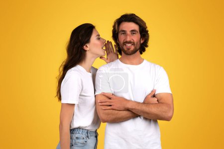 Téléchargez les photos : Homme caucasien heureux confiant avec les bras croisés souriant tandis qu'une femme murmure dans son oreille, les deux vêtus de chemises blanches sur un fond jaune vif, studio. Potins, nouvelles - en image libre de droit