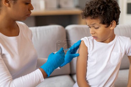 Téléchargez les photos : Médecin noir femme en gants de protection faisant la vaccination pour le petit garçon à l'intérieur de la maison, gros plan de l'enfant afro-américain mâle effrayé se faire vacciner contre diverses maladies, gros plan - en image libre de droit