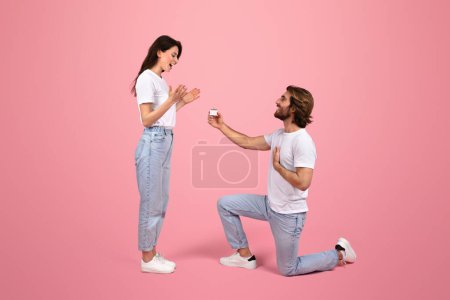 Die überglückliche kaukasische junge Frau reagiert überrascht, als der Mann mit einem Verlobungsring kniet, sowohl in lässigen weißen Oberteilen als auch in Jeans vor sanftem rosa Hintergrund, Studio