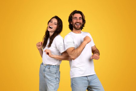 Photo for Joyful and lively glad millennial caucasian couple dancing and laughing, with the woman looking at the man, both in casual white t-shirts, having fun on a yellow background, studio - Royalty Free Image