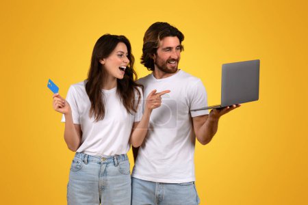 Photo for Excited surprised happy caucasian young woman holding a credit card and pointing at laptop screen shown by a smiling man, both in white t-shirts, against a yellow backdrop, studio - Royalty Free Image