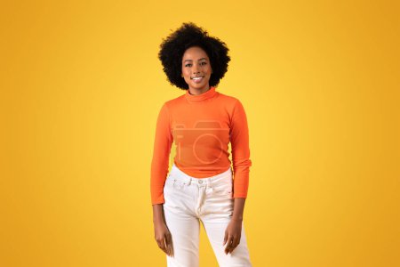 Téléchargez les photos : Une jeune femme noire joyeuse avec un sourire radieux se tient avec confiance, portant un col roulé orange vif et un pantalon blanc sur un fond jaune vif, studio. Mode de vie, étude - en image libre de droit
