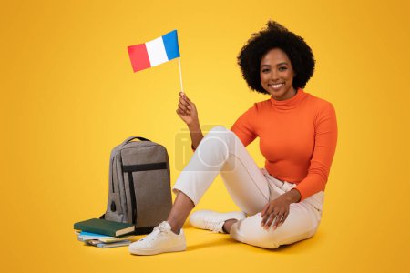 Foto de Sonriente mujer afroamericana milenaria con el pelo rizado sentado con las piernas cruzadas en el suelo, sosteniendo una bandera francesa, con una mochila y libros cerca, sobre un fondo amarillo - Imagen libre de derechos