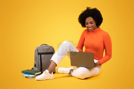 Photo for Engaged millennial African American student with curly hair using a laptop, sitting cross-legged on the floor, with a grey backpack and books beside her, on a yellow background - Royalty Free Image