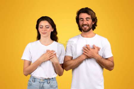 Photo for Grateful european young couple with eyes closed and hands on heart, feeling thankful and serene in plain white t-shirts, standing against a vivid yellow background, studio - Royalty Free Image