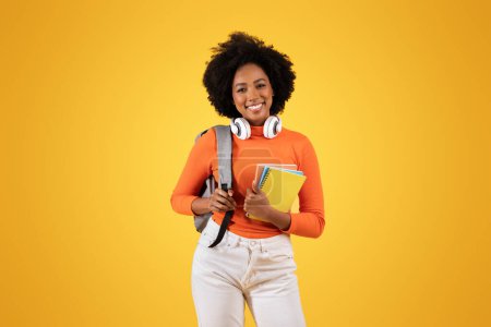 Téléchargez les photos : Jeune femme noire souriante avec un casque autour du cou tenant des livres et portant une chemise orange avec un sac à dos gris sur fond jaune, studio. Éducation, études - en image libre de droit