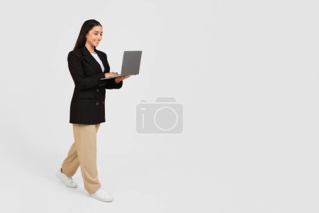 Téléchargez les photos : Femme d'affaires joyeuse en tenue formelle marchant et travaillant sur son ordinateur portable contre un fond studio propre et gris clair, espace libre - en image libre de droit