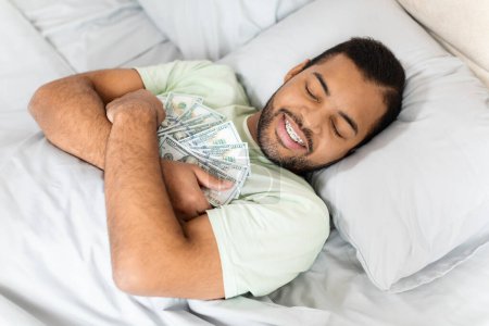 Foto de Feliz joven negro abrazando dinero en efectivo mientras duerme, acostado en la cama. Vista superior de sonriente hombre afroamericano abrazando billetes de dólar, mantener sus ahorros con él - Imagen libre de derechos