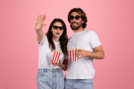 Téléchargez les photos : Couple enthousiaste portant des lunettes 3D, tendu excitamment vers quelque chose d'invisible tout en tenant des boîtes de pop-corn à rayures rouges et blanches, sur fond rose, véhiculant un thème de soirée cinéma - en image libre de droit