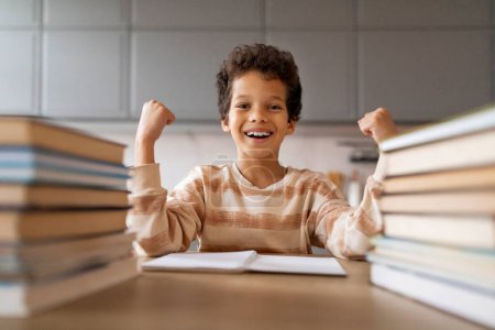 Fröhlicher schwarzer Schuljunge, der zu Hause eine erfolgreiche Lerneinheit feiert, glückliches männliches Kind mit erhobenen Fäusten und strahlendem Lächeln, das neben einem Stapel Bücher sitzt und nach getaner Arbeit Freude empfindet