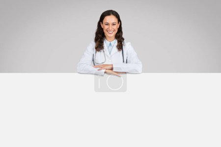 Photo for Cheerful european lady doctor in white uniform, leaning on huge white blank placard for medical advertising offers, against grey background, free space - Royalty Free Image