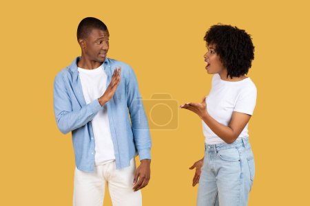 A black man defensively gesturing stop with his hand as a woman animatedly talks to him, both in casual clothes on a mustard yellow background. Relationship problems, quarrel, scandal
