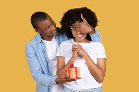 Freudig freudiger schwarzer junger Mann überrascht seine entzückte Freundin mit einem kleinen Geschenk mit roter Schleife und verdeckt spielerisch ihre Augen für eine Überraschung vor gelbem Hintergrund