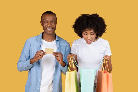 Foto de Emocionado sonriente pareja afroamericana adictos a las compras con coloridas bolsas de compras y una tarjeta de crédito, sonriendo felizmente a sus compras exitosas sobre un fondo amarillo sólido, estudio - Imagen libre de derechos