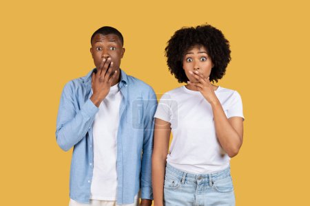 Photo for Shocked black young couple in casual attire with wide eyes and hands over their mouths expressing surprise against a solid mustard yellow backdrop, studio. Sale, surprise - Royalty Free Image