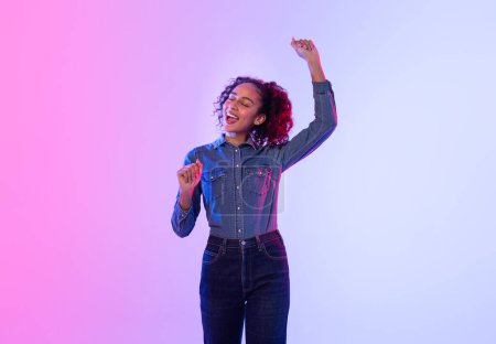 Téléchargez les photos : Femme noire exubérante aux cheveux bouclés dansant joyeusement, portant une tenue denim complète sur fond de dégradé rose vif à bleu, exprimant bonheur et liberté - en image libre de droit