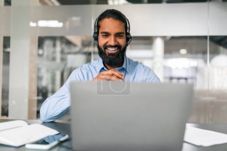 Foto de Retrato de un positivo barbudo milenario indio empleado de servicio al cliente que trabaja en la oficina, usando auriculares, mirando la pantalla de la computadora portátil y sonriendo. Concepto de trabajadores - Imagen libre de derechos