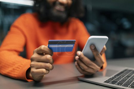 Photo for Cropped of bearded eastern man using phone and credit card while working on laptop at office. Hands of businessman paying for goods and services online, shopping or banking - Royalty Free Image