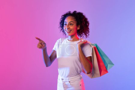 Téléchargez les photos : Femme noire exubérante aux cheveux bouclés, souriante et pointant vers le côté, portant des sacs à provisions vibrants, sur fond de dégradé rose fluo et bleu, suggérant une vente ou une offre festive - en image libre de droit