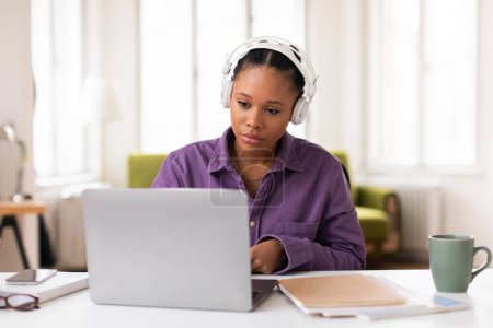 Photo for African american female student wearing headphones deeply focused on her studies while working on laptop at bright home office setting, free space - Royalty Free Image