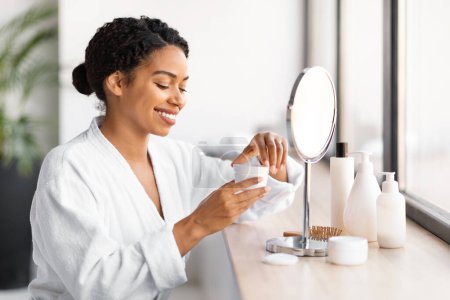 Foto de Mujer negra sonriente en bata blanca abriendo tarro de crema facial, hembra afroamericana sonriente preparándose para su rutina de cuidado de la piel en casa, sentada cerca del espejo en el baño, primer plano - Imagen libre de derechos