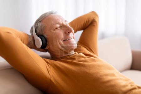 Téléchargez les photos : Homme âgé détendu jouissant d'une bonne musique à la maison, allongé sur le canapé avec les yeux fermés et les bras derrière la tête, utilisant un casque sans fil, écoutant des chansons préférées, gros plan - en image libre de droit