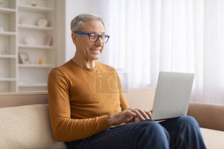 Photo for Positive grandfather wearing eyeglasses sitting on couch with laptop computer on his lap, websurfing, reading news online. Elderly man working online from home. Remote job for seniors - Royalty Free Image