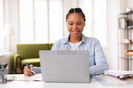 Foto de Alegre estudiante negra con la pluma en la mano sonriendo mientras mira su computadora portátil, lo que sugiere una agradable experiencia de aprendizaje en línea, sentado en el escritorio en casa - Imagen libre de derechos