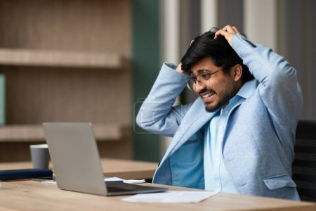 Photo for Unhappy Indian professional businessman grasping his head in frustration while staring at his laptop, looking at mirror and mistake in modern office. Workplace stress and panic, business failure - Royalty Free Image