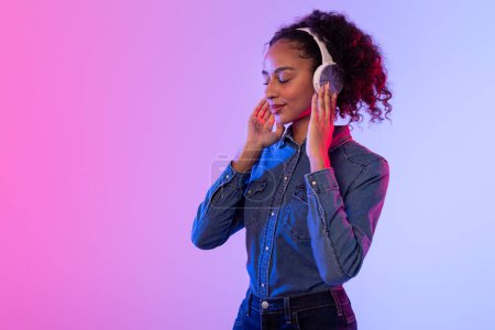 Téléchargez les photos : Contenu dame noire avec les yeux fermés savourant le son, portant des écouteurs en argent et veste en denim, sur fond dégradé rose et bleu lumineux - en image libre de droit