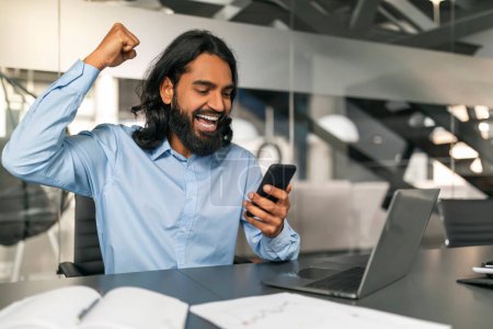Photo for Excited indian guy manager looking at smartphone screen, gesturing raising hand up and screaming, have great news, celebrating business success, modern office interior, copy space - Royalty Free Image