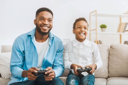 Téléchargez les photos : Happy afro papa profiter du jeu vidéo avec sa fille mignonne, jouer avec des joysticks à la maison, espace libre - en image libre de droit