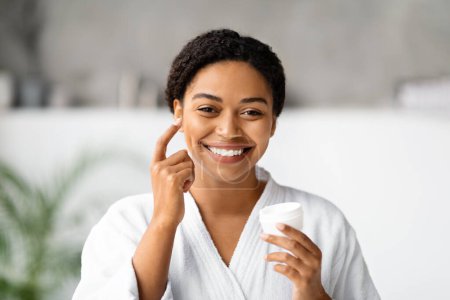 Téléchargez les photos : Femme noire joyeuse appliquant une crème hydratante sur sa joue, heureuse femme afro-américaine en peignoir blanc tenant le pot du produit et souriant à la caméra, faisant une routine de beauté à la maison - en image libre de droit