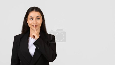 Smiling young businesswoman in black suit making quiet sign with her finger to her lips, looking aside at free space against light grey background, panorama