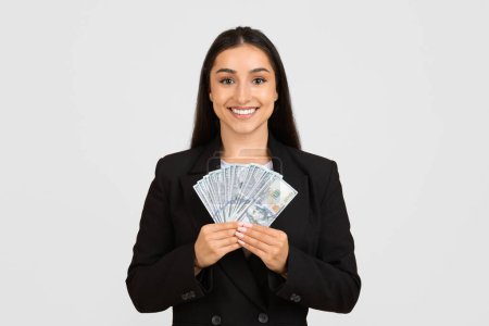 Fröhliche Geschäftsfrau im schwarzen Anzug zeigt stolz Fan von Hundert-Dollar-Scheinen mit selbstbewusstem Lächeln und posiert vor hellgrauem Studiohintergrund