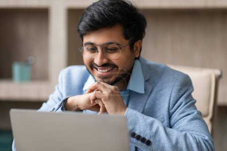 Photo for Young Hindu professional businessman wearing eyeglasses looking at laptop, smiles while browsing internet at modern workplace indoor. Entrepreneur using computer in office - Royalty Free Image