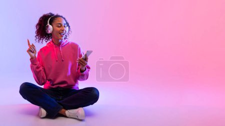 Téléchargez les photos : Joyeux dame noire portant sweat à capuche rose et jeans, écoute de la musique sur écouteurs blancs et utilise smartphone, assis jambes croisées sur le sol contre dégradé rose toile de fond, espace libre - en image libre de droit