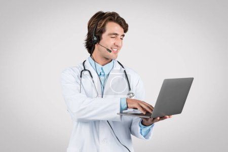 Cheerful male doctor wearing headset and lab coat while expertly navigating telehealth platforms on laptop, showcasing modern healthcare communication