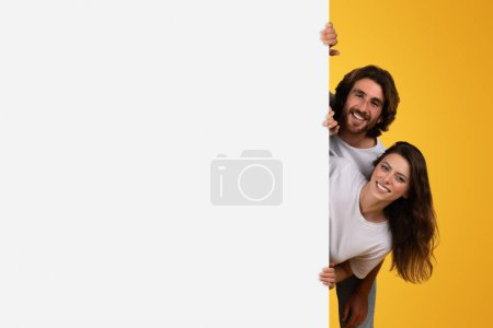 Joyous european couple playfully peeking around a blank white space for copy or advertisement, with a vibrant yellow backdrop, engaging with a bright and cheerful demeanor