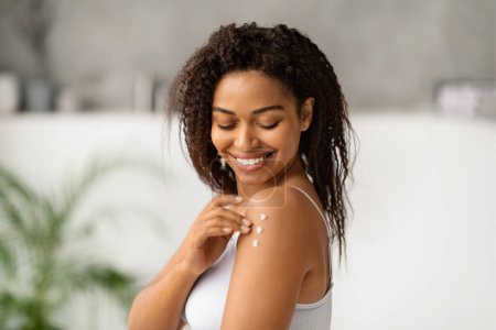 Foto de Mujer negra alegre sonriendo mientras se aplica loción corporal en el hombro, joven mujer afroamericana feliz disfrutando de su rutina de cuidado de la piel, de pie en un baño moderno y luminoso, espacio para copiar - Imagen libre de derechos