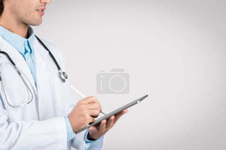 Side view of concentrated male doctor in white coat with stethoscope, using digital tablet to record patient data or consult medical records, free space