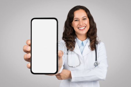 Photo for Telemedicine offer. Young woman doctor showing smartphone with white blank screen, presenting mockup for app or ad, standing over grey studio background. Medicine and mobile technology - Royalty Free Image