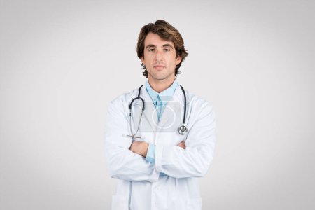 Professional male doctor with wavy hair, wearing white lab coat and stethoscope, stands confidently with arms crossed against grey background, conveying expertise and trustworthiness