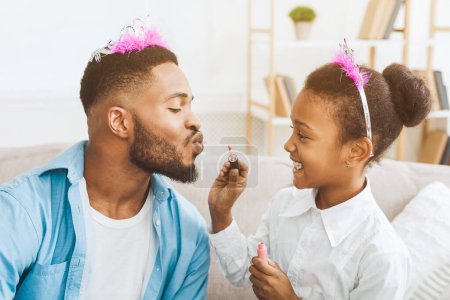 Téléchargez les photos : Petite fille appliquant du rouge à lèvres sur les lèvres des pères, s'amusant à la maison - en image libre de droit