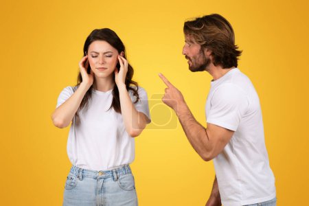 Gestresste Europäerin, die ihre Ohren bedeckt, während ein Mann wütend mit dem Finger auf sie zeigt und Konflikte oder Meinungsverschiedenheiten darstellt, beide tragen weiße Oberteile auf gelbem Hintergrund, Studio