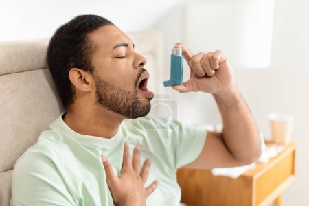 Photo for Handsome african american man sitting alone in bed at home and using an asthma pump. Black Guy Inhaling Asthmatic Cure at Home, using medical inhaler to prevent shortness of breath - Royalty Free Image
